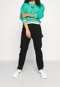 Persona in felpa turchese con logo colorato, pantaloni neri e sneakers bianche, in piedi contro uno sfondo bianco con le braccia incrociate.