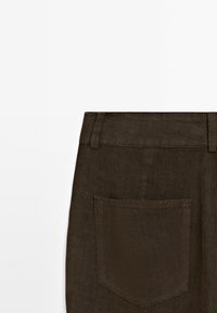 Vue arrière d'un pantalon marron foncé montrant la ceinture avec passants et une poche arrière rectangulaire unique sur fond blanc.