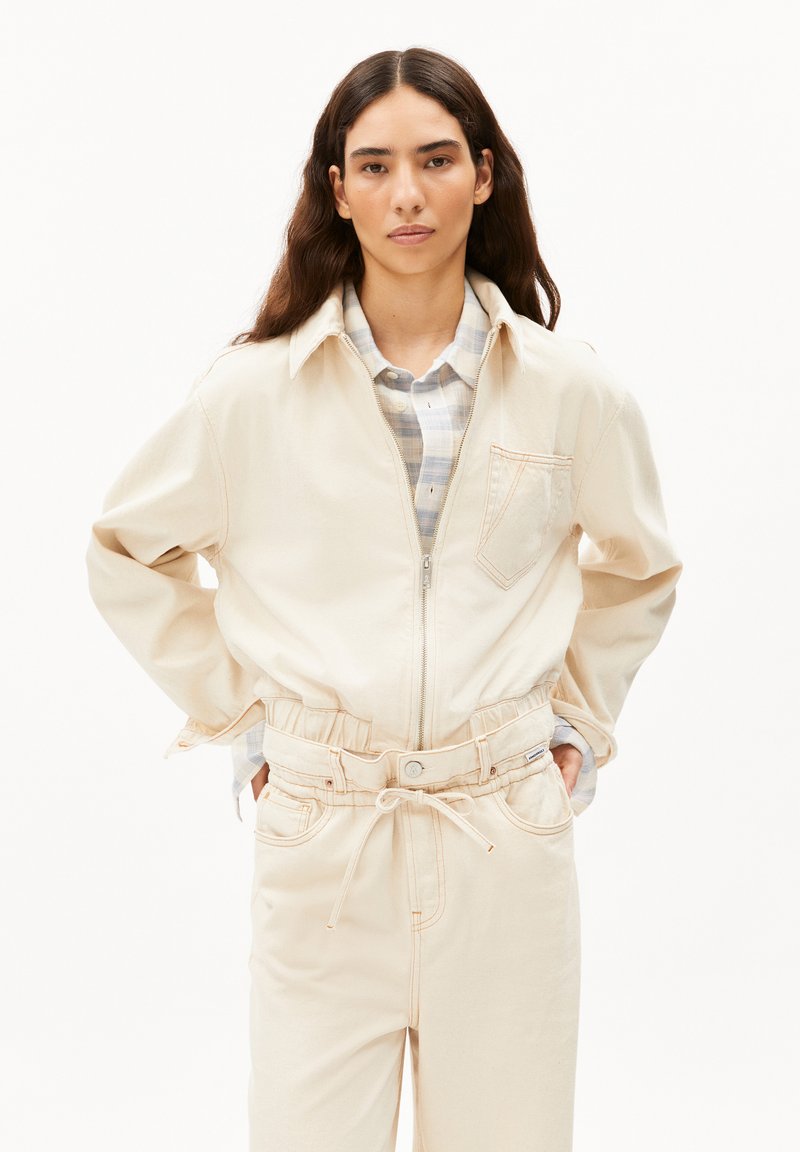 Hellbeige Zip-Jacke mit Kragen, oversized Schnitt und einer Brusttasche. Kombiniert mit passenden, schmal zulaufenden Hosen und einem karierten Hemd.