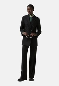 Black double-breasted blazer with a green inner layer and wide-legged black trousers. Model holds sunglasses, styled with a patterned neck scarf.