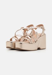 Sandali platform metallici oro con fasce incrociate e cinturino alla caviglia regolabile. Tacco a zeppa spesso con una texture liscia e design della punta arrotondato.