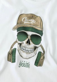 Bílé tričko s grafikou lebky, která má na sobě zelené sluneční brýle a sluchátka, s béžovou kšiltovkou s nápisem "Chicago" a textem "URBAN YOUTH."