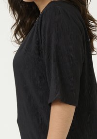 Femme portant un haut noir texturé à manches courtes, montrée de côté avec des cheveux foncés ondulés sur un fond neutre.