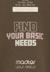 Camiseta de algodón marrón con texto llamativo, que incluye "ENCUENTRA TUS NECESIDADES BÁSICAS" y "domina tus habilidades". Estilos y tamaños de fuente variados.