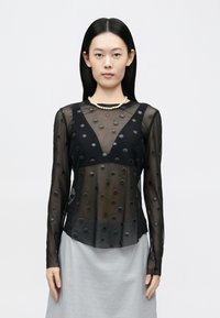 Čierna priehľadná blúzka s dlhým rukávom s vystúpenou bodkovanou textúrou. Výstrih do V, zdobené rukávy, v kombinácii s svetlosivou sukňou.