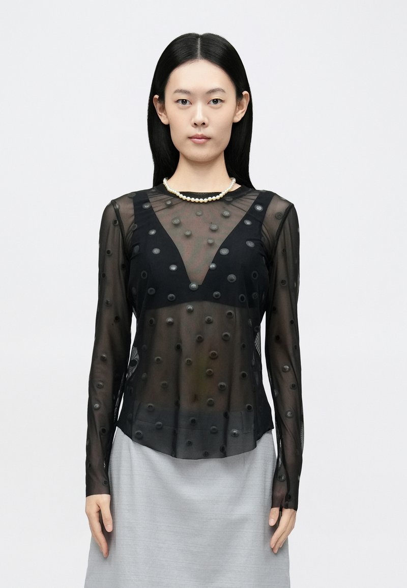 Čierna priehľadná blúzka s dlhým rukávom s vystúpenou bodkovanou textúrou. Výstrih do V, zdobené rukávy, v kombinácii s svetlosivou sukňou.