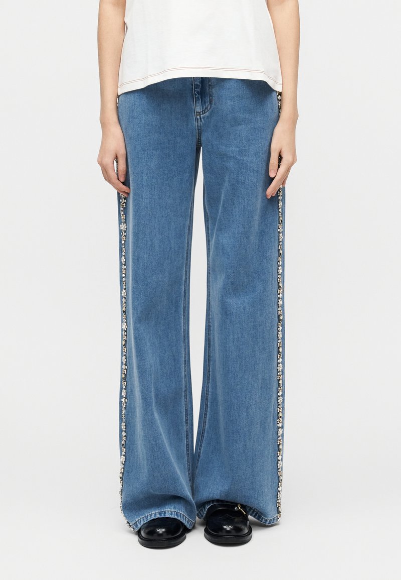 Άτομο που φοράει φαρδιά μπλε τζιν με διακοσμητική τρέσα από παγιέτες στις πλευρικές ραφές, λευκή μπλούζα και μαύρα παπούτσια, στέκεται μπροστά σε απλό λευκό φόντο.