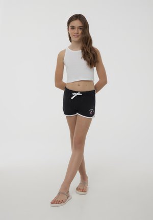 Pantaloni sportivi neri con bordi bianchi, design ricamato, abbinati a un top corto bianco. Calzature: sandali piatti argentati. Tessuto liscio.