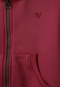 Sudadera con cremallera burdeos hecha de tela suave, con un pequeño corazón bordado y bolsillos frontales con puños acanalados.