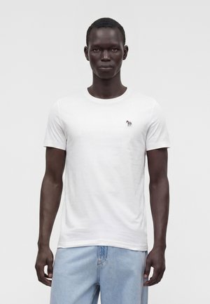 Bărbat care poartă un tricou alb simplu cu mâneci scurte și un mic logo cu zebră pe piept și blugi albastru deschis, stând în fața unui fundal alb.