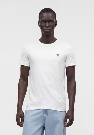 Homme portant un t-shirt blanc uni à manches courtes avec un petit logo zèbre sur la poitrine et un jean bleu clair, debout devant un fond blanc.