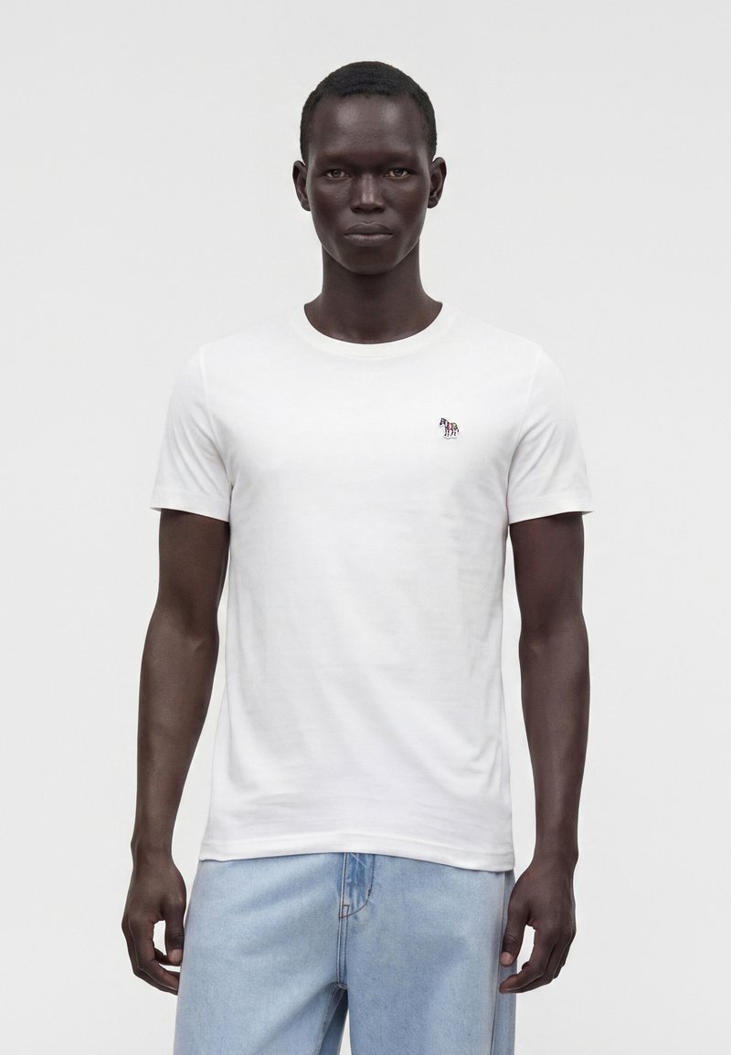 Άνδρας που φοράει ένα απλό λευκό t-shirt με κοντά μανίκια και μικρό λογότυπο ζέβρας στο στήθος και ανοιχτόχρωμα μπλε τζιν, στέκεται μπροστά σε λευκό φόντο.