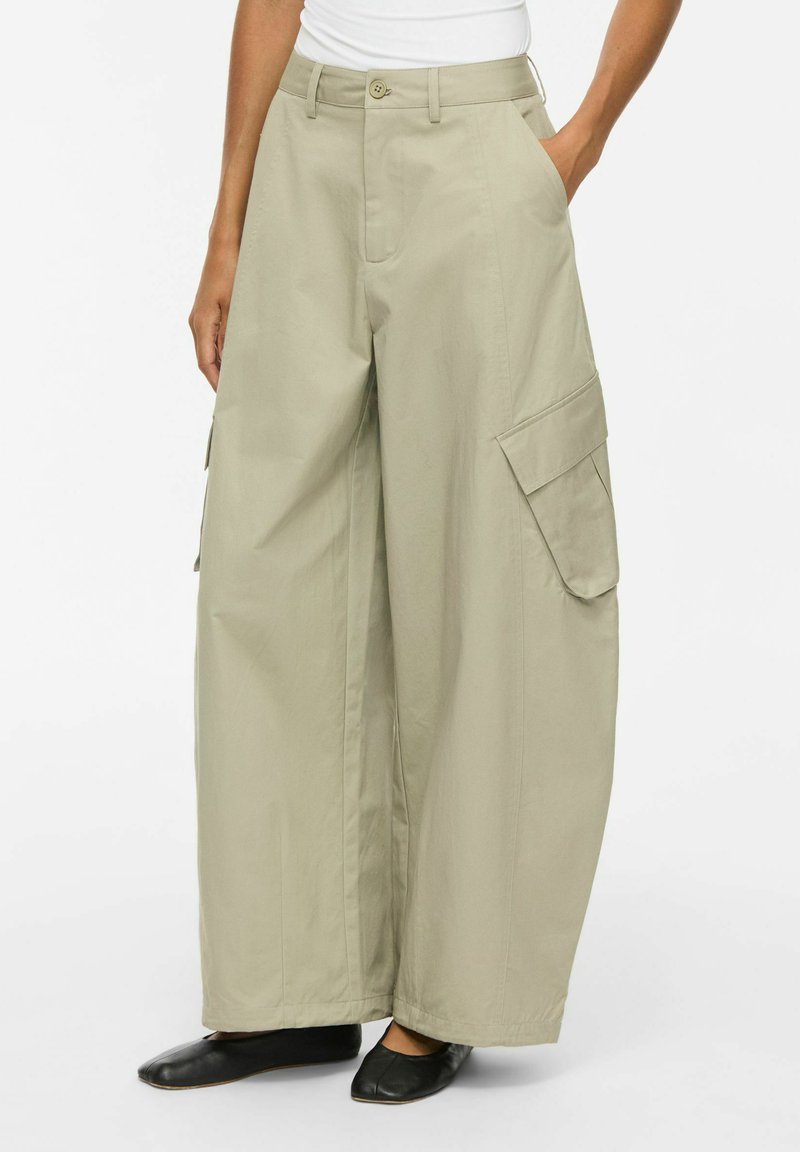 Pantalon cargo beige à jambe large avec grandes poches latérales, porté avec des chaussures plates noires et un haut blanc rentré, montré de la taille aux pieds.