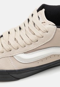 Baskets beige et noires avec bande latérale blanche, lacets beiges épais, dessus en mesh et daim, et doublure intérieure noire.