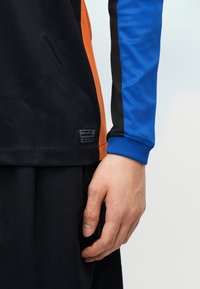 Δεξί χέρι ατόμου που φοράει μακρυμάνικο αθλητικό μπλουζάκι σε μαύρο, πορτοκαλί και μπλε χρώμα, με μια μικρή ορθογώνια ετικέτα κοντά στο τελείωμα.