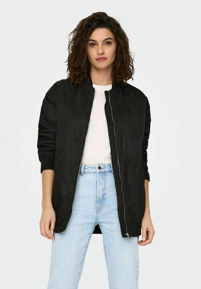 Veste bomber noire avec fermeture éclair à l'avant, poignets côtelés et texture lisse et brillante, assortie à une chemise blanche et un jean bleu clair.
