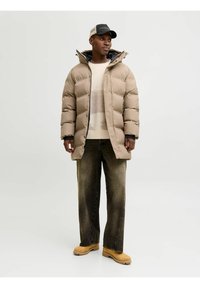 Chaqueta de plumas beige con un alto collar, capucha y puños elásticos. Lucida sobre un suéter crema con estampado, combinada con jeans oscuros y botas de color marrón claro.