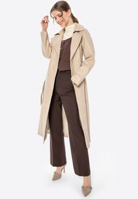Trench coat beige chiaro con un ampio colletto, abbinato a un top aderente marrone e pantaloni a gamba larga. La modella si erge con un sorriso, tenendo i capelli.
