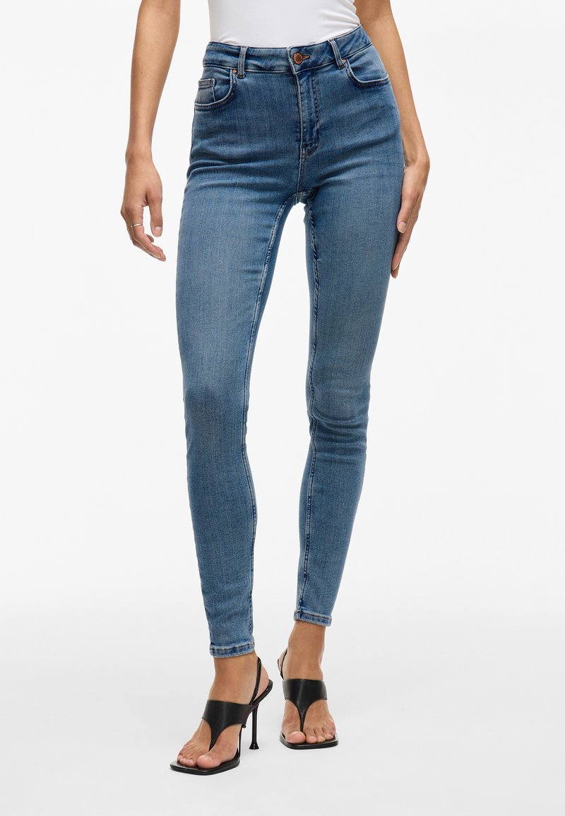 Femme portant un jean slim bleu taille haute et des sandales noires à talons ouverts sur fond blanc.