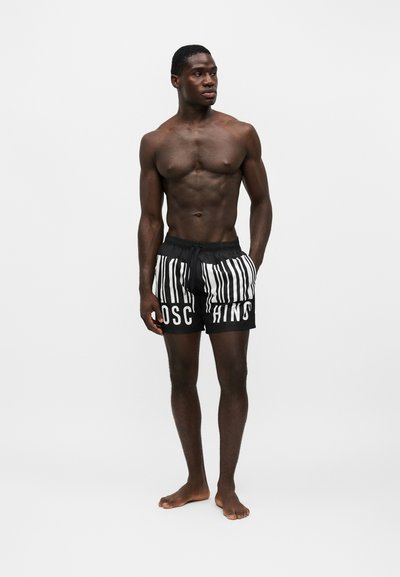 Homme musclé debout pieds nus portant un short de bain noir avec un motif code-barres blanc et un texte blanc partiel à l'avant.