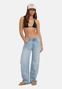 Μαύρο μπικίνι τοπ, ανοιχτό μπλε φαρδύ τζιν παντελόνι, καφέ σάνταλ και μπεζ καπέλο με λογότυπο. Το μοντέλο ποζάρει με το ένα χέρι στην τσέπη.