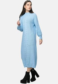 Kvinde iført en lyseblå midi sweaterkjole med kabelstrik og ballonærmer samt sorte platformsstøvler, poserende på en hvid baggrund.