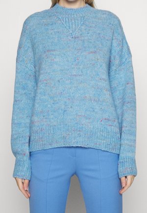 Personne portant un pull en tricot bleu clair moucheté avec des poignets et un ourlet côtelés, associé à un pantalon bleu uni, visible du cou aux hanches.