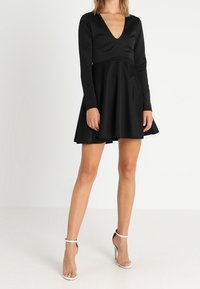 Musta pitkähiha dressi, jossa syvä V-kaula-aukko ja levenevä helma. Valmistettu sileästä materiaalista, jossa vartalonmyötäinen yläosa ja minimalistinen muotoilu.