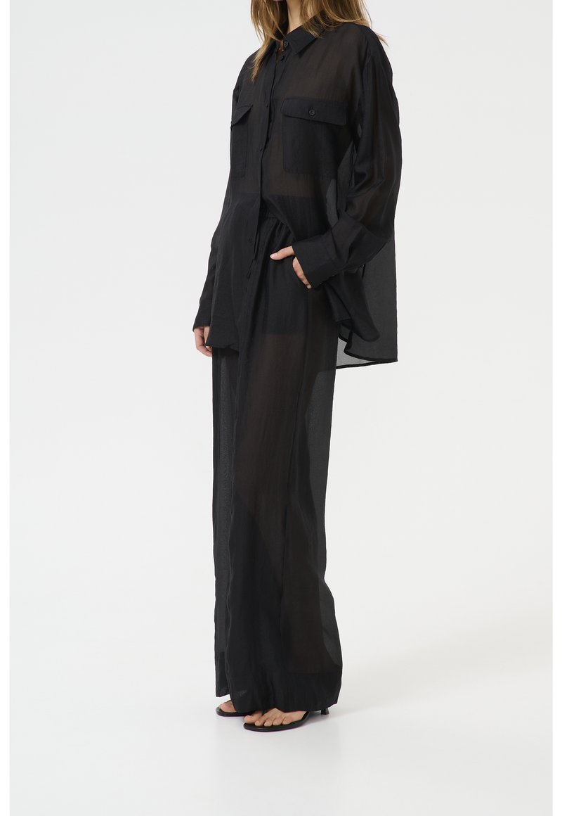 Chemise noire, transparente,  à manches longues avec deux poches poitrine, coupe décontractée et ourlet arrondi, associée à un pantalon noir taille haute.