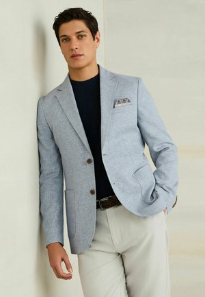 Man draagt een lichtblauwe blazer, marineblauwe trui, beige broek en een patroon zakdoek, leunend tegen een muur met één hand in de zak.