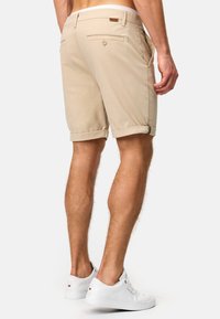 Beige shorts med opbrettede manchetter, for- og baglommer, lavet af et struktureret stof. Kombinationen med hvide sneakers. Etiketten er synlig på taljebåndet.