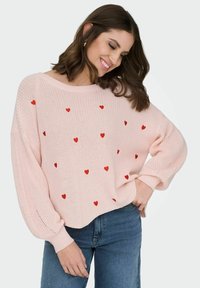 Femme souriante portant un pull en tricot rose clair avec des cœurs rouges, associé à un jean bleu, posant devant un fond uni.