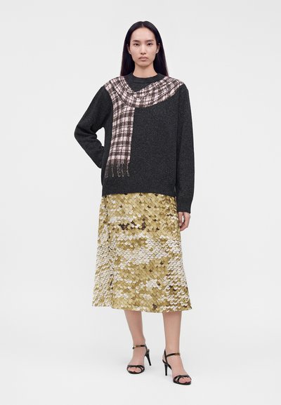 Femme portant un pull noir à motif à carreaux roses et une jupe midi à motifs dorés et blancs, debout, une main sur la hanche et des sandales noires.