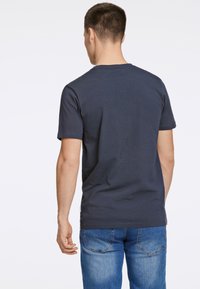 Tummansininen puuvillapaita, jossa lyhyet hihat ja pyöreä kaula-aukko, kuvattu takapuolelta. Yhdistetty vaaleansinisiin farkkuihin.