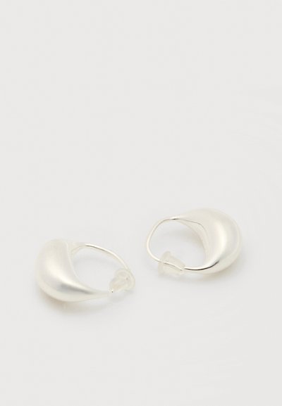 Καμπυλωτά ασημένια σκουλαρίκια κρίκοι με λεία επιφάνεια και διαφανή πλαστικά πίσω μέρη, τοποθετημένα πάνω σε λευκή επιφάνεια.