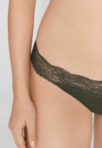 Dunkelgrüner String mit Spitzenbesatz und glattem Stoffkörper sowie dekorativem Spitzenbund. Nahaufnahme, die Textur und Details hervorhebt.