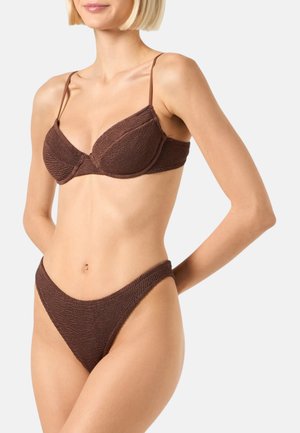 Vrouw draagt een gestructureerde bruine tweedelige bikini met dunne bandjes boven en hoog uitgesneden broekjes, staand tegen een effen achtergrond.