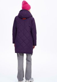 Fialový prošívaný dlouhý kabát s kapucí, s výrazným oranžovým detailem na zadní straně. Vytvořený v kombinaci se šedými rozšířenými džíny a teniskami.