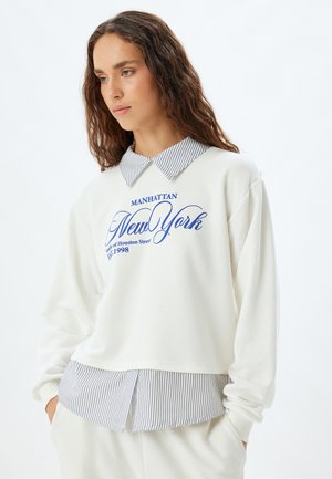 Sudadera blanca con cuello y puños a rayas, con el texto azul "Manhattan New York" y un corte relajado. Textura más suave con un diseño casual.