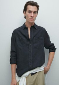 Mörk denim skjorta med button-down krage, två bröstfickor och uppvikta ärmar. I kombination med en knuten vit tröja och ljusa byxor.