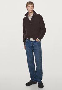 Ciemnobrązowy sweter z ribbowanym zamkiem, jasna koszula z kołnierzykiem pod spodem, niebieskie dżinsy i czarne buty. Model stoi na tle białej ściany.