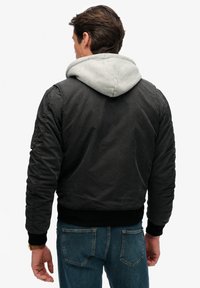 Sort bomberjakke med grå hætte, elastiske manchetter og linning; har en glat tekstur og syningsdetaljer på ærmerne.