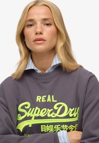 Grijze sweatshirt met neon groene "SuperDry" tekst, met een zachte textuur en ronde halslijn, gelaagd over een blauw-wit gestreept shirt.