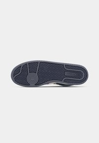 Dark rubber shoe sole with zigzag grip pattern, featuring branding and a circular accent. The edge is textured with a smooth finish.
