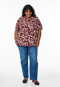 Camisa de manga corta abotonada con un patrón floral en rojo y crema, combinada con unos vaqueros rectos azul y zapatos sin cordones oscuros de tejido.