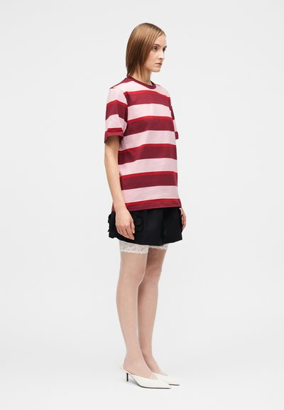 Kobieta w czerwono-różowej koszulce w paski na krótki rękaw, czarnej spódnicy z kwiatowymi detalami, białych koronkowych rajstopach i białych klapkach na obcasie, stojąca bokiem.