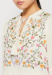 Broderad krämfärgad blus med blommönster i rosa, orange och blått, strukturerad tyg och en halsringning med knytning och fransar.