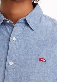 Jasnoniebieska koszula dżinsowa z kołnierzykiem zapinanym na guziki, białymi guzikami i małym czerwonym logo Levi's na piersi. Miękka tekstura i swobodny design.