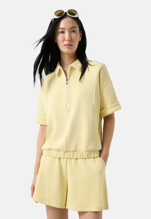 Junge Frau mit langen schwarzen Haaren, die ein hellgelbes kurzärmeliges Zip-up-Top und passende Shorts trägt, mit Sonnenbrille auf dem Kopf.