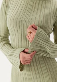 Robe en tricot côtelé à manches longues de couleur vert pâle, avec une texture douce, des poignets évasés larges et un décolleté arrondi. Anneau en or à la main.